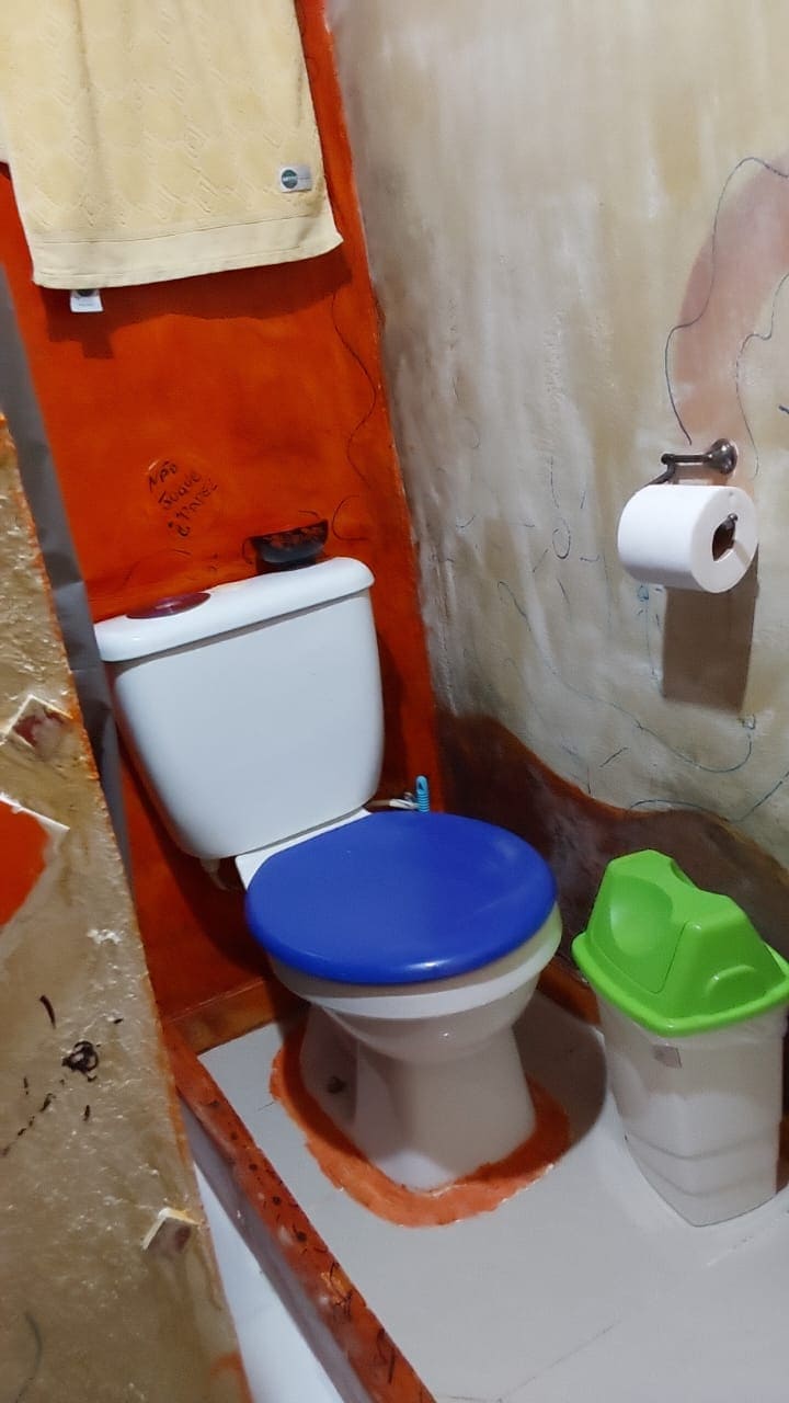 The bathroom features a compact space with an orange textured wall. A toilet with a blue seat is positioned next to a green waste bin. A roll of toilet paper is neatly placed on a wall-mounted holder. A yellow towel hangs above, adding a touch of color.