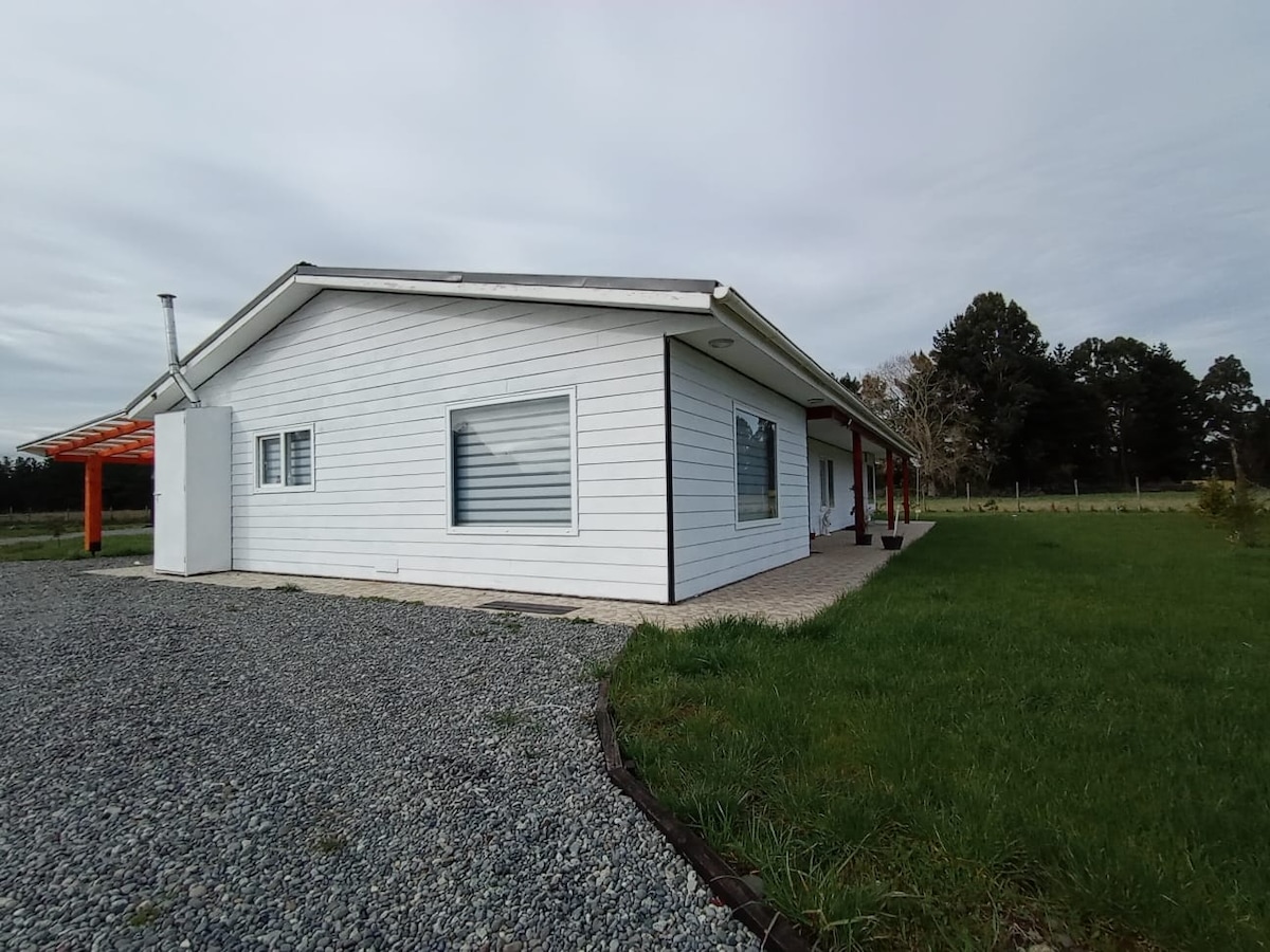 The exterior of the one-story house is showcased, featuring a white facade with large windows and a sloped roof. A stone pathway leads to the entrance, while a grassy area and gravel driveway surround the home, providing an open, inviting space.