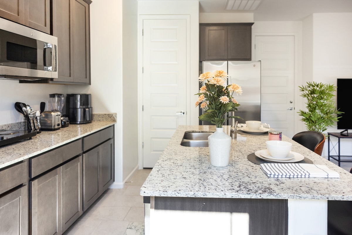 The kitchen area features contemporary cabinetry and a large granite island with seating. Equipped with stainless steel appliances, including a microwave and coffee maker, the space is well-lit by natural light. Stylish decor includes a vase of flowers and neatly arranged dining essentials.