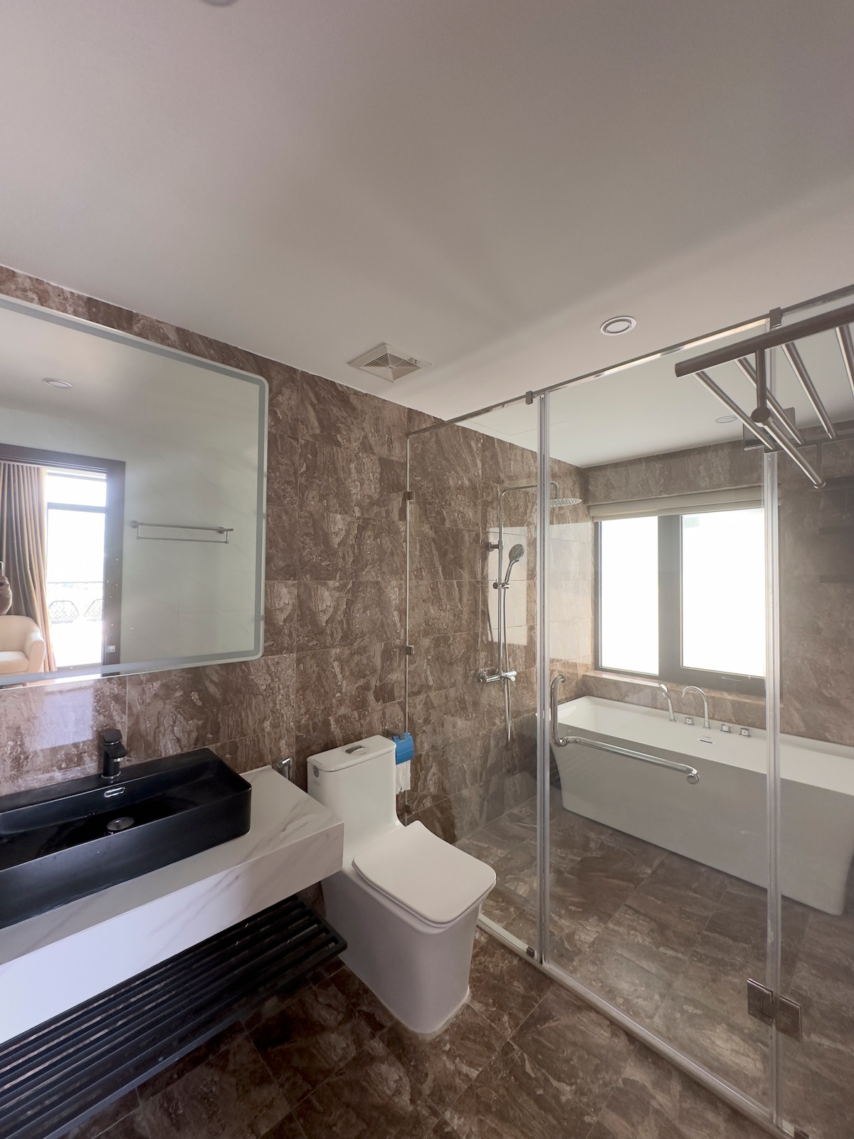 A modern bathroom features a sleek dark countertop with a vessel sink. A glass-enclosed shower with fixtures is adjacent to a freestanding bathtub. Large windows provide natural light, framing the view outside. Neutral stone tiles create a cohesive and polished look.