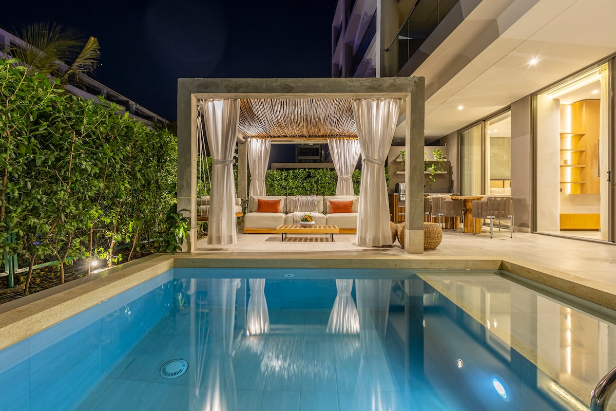 A private terrace features a lounge area with a thatched roof, surrounded by lush greenery. The inviting pool reflects the soft glow of exterior lighting, enhancing the serene atmosphere. Large glass doors provide a seamless connection to the indoor living space.