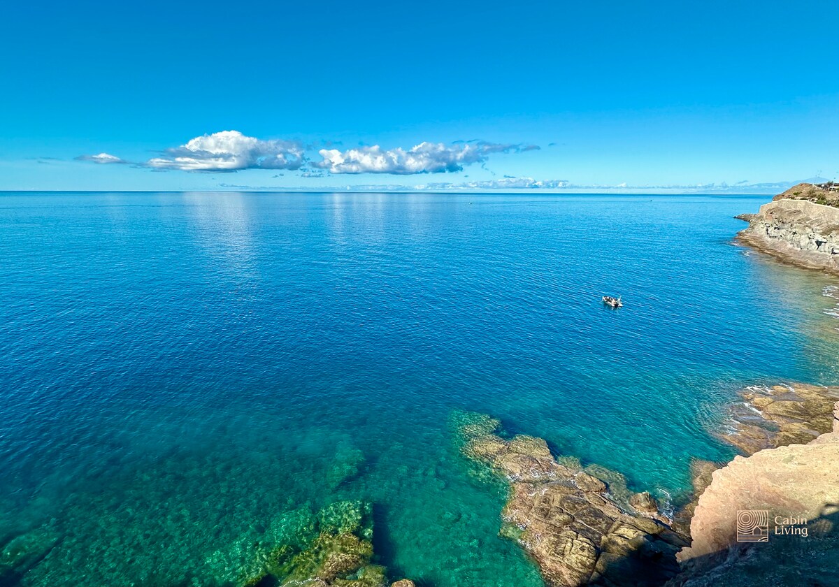 A clear, tranquil sea stretches to the horizon, displaying vibrant turquoise and deep blue tones. A rocky shoreline is visible in the foreground, complementing the serene water. A small boat can be seen in the distance, enhancing the sense of peacefulness and connection to nature.