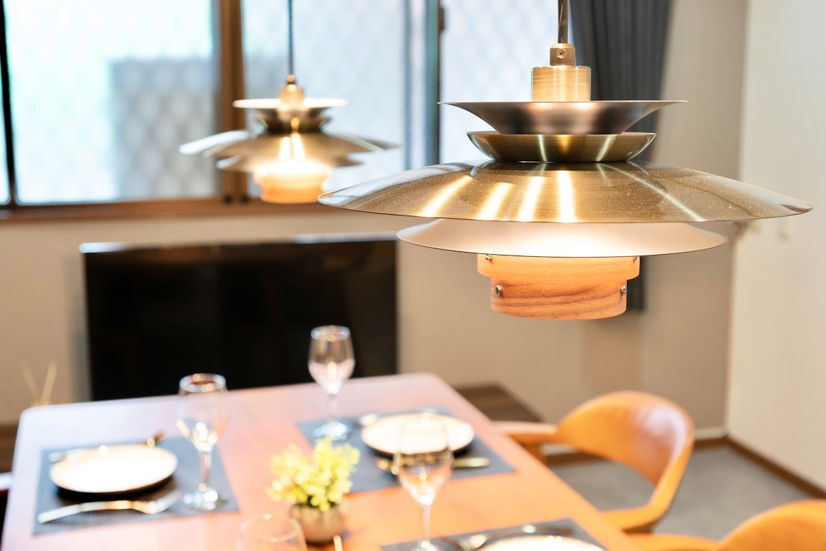 A dining area is seen featuring a wooden table set with plates and glassware. Two modern pendant lights hang above, casting a warm glow. A minimalist design is complemented by soft furnishings and natural light filtering through the windows.