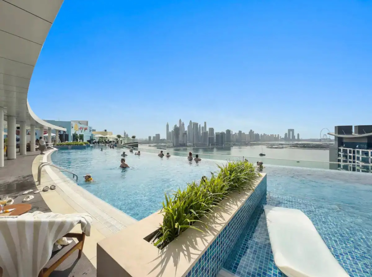 An elevated infinity pool is shown with sleek edges, overlooking the scenic skyline of Palm Jumeirah. Visitors are seen enjoying the water, surrounded by lush greenery and modern lounge chairs. Clear blue skies complement the stunning waterfront views.