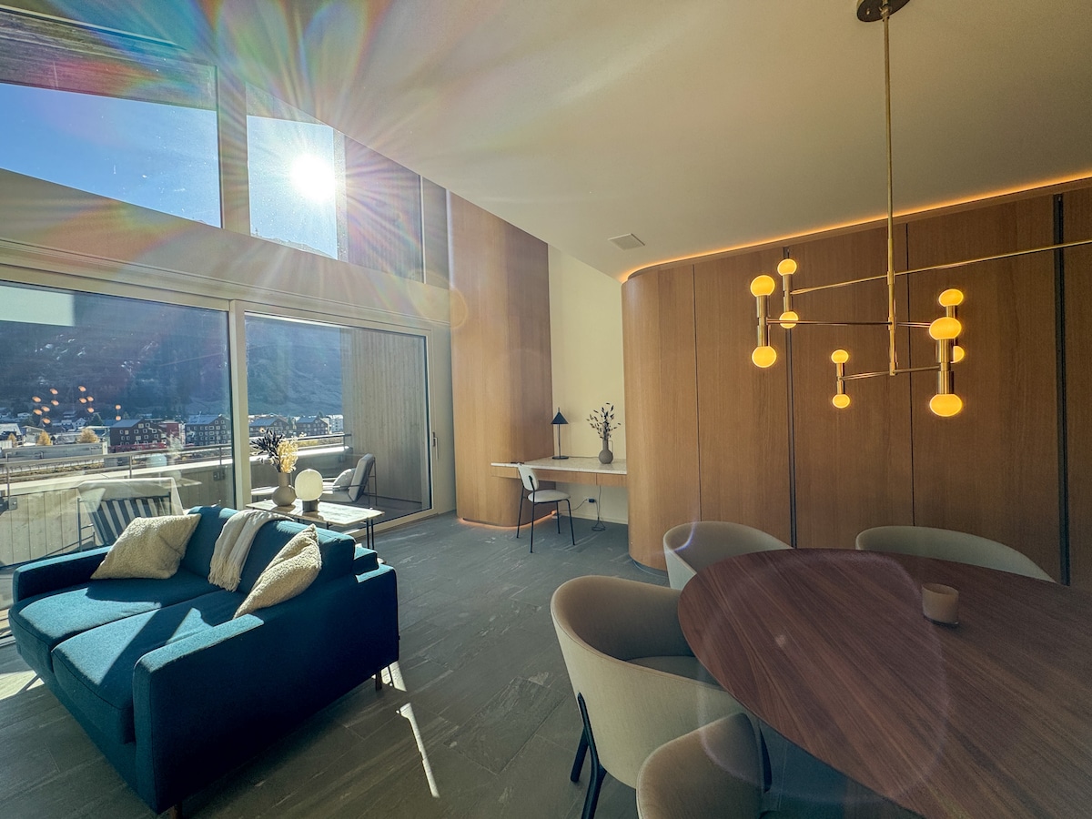 A modern living area features a sleek blue sofa and a round dining table surrounded by light-colored chairs. Sunlight streams through expansive windows, illuminating warm wood paneling and a minimalist workspace with a desk. A warm, inviting ambiance is created by stylish lighting.