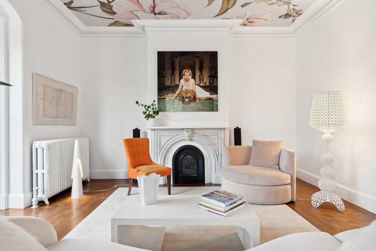 A light-filled living area features a circular couch and a single orange chair, complementing the elegant marble fireplace. Above, a colorful floral ceiling design adds character. A white rug anchors the space, with a modern lamp and stylish decor enhancing the sophisticated interior.