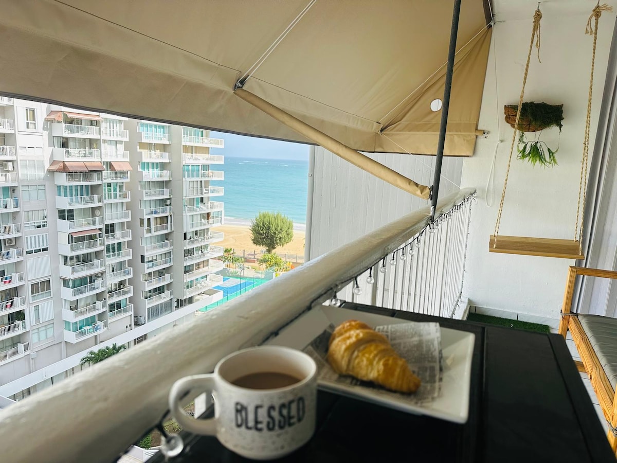 A balcony offers scenic views of the beach and ocean. A small table holds a coffee cup and a croissant. A cozy swing chair hangs nearby, and greenery adds a touch of nature to the space. The building's exterior features a light-colored facade.
