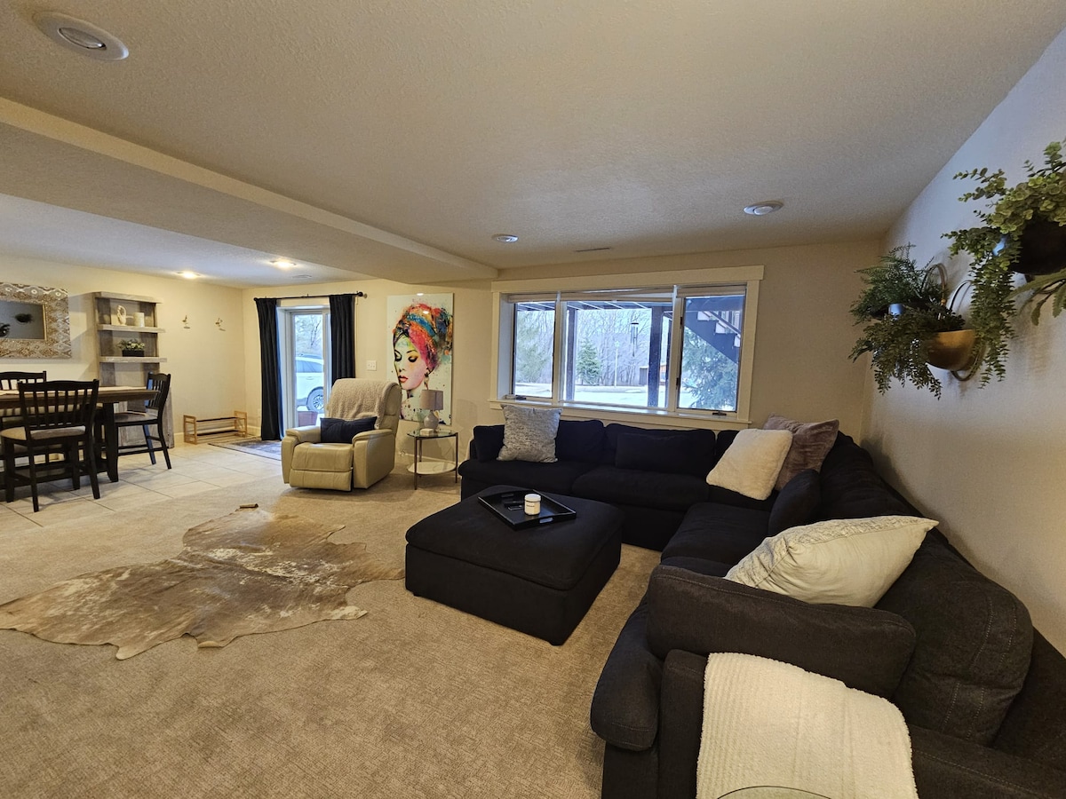 A spacious living area features a large sectional sofa in dark fabric, a cozy recliner, and a wooden dining table with chairs. Natural light enters through multiple windows, illuminating the soft carpet and decorative elements, including plants on the wall and a throw blanket on the sofa.