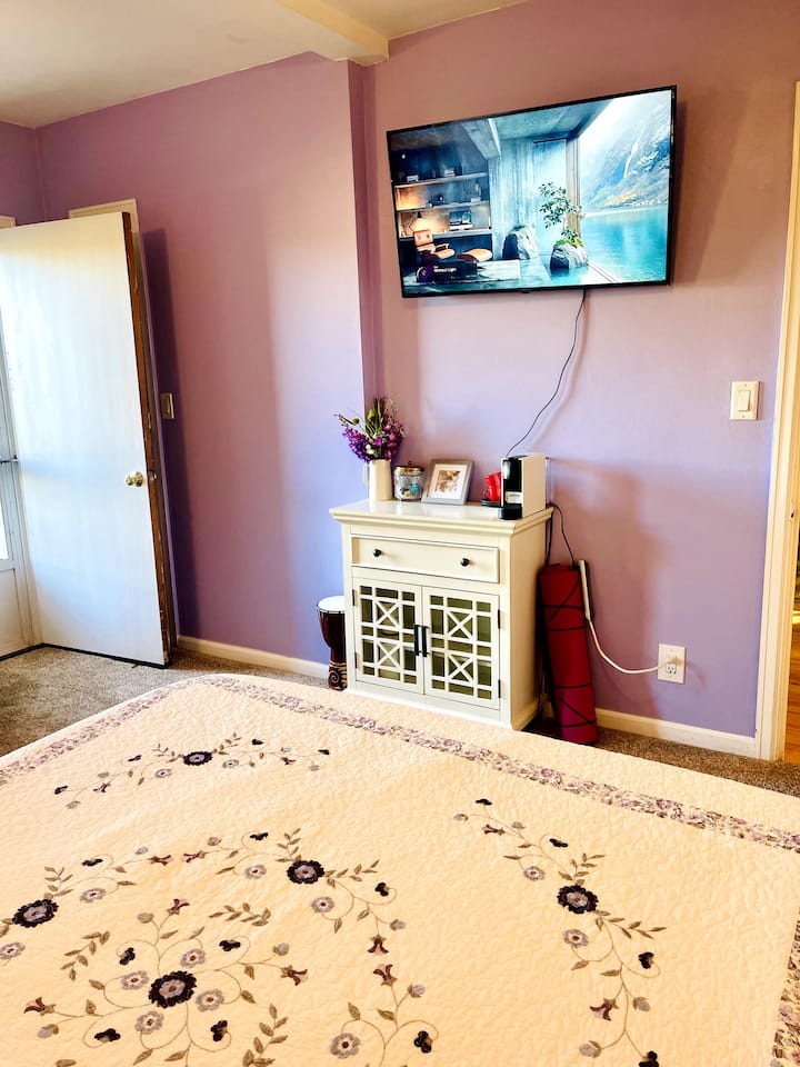 منظر غرفة نوم لافندر من سرير بحجم كبير يعمل عن بعد. يعرض تلفزيون إل جي الذكي وبوفيه ونسبريسو وسجادة يوغا وأسطوانة إندونيسية مصنوعة يدويًا. 