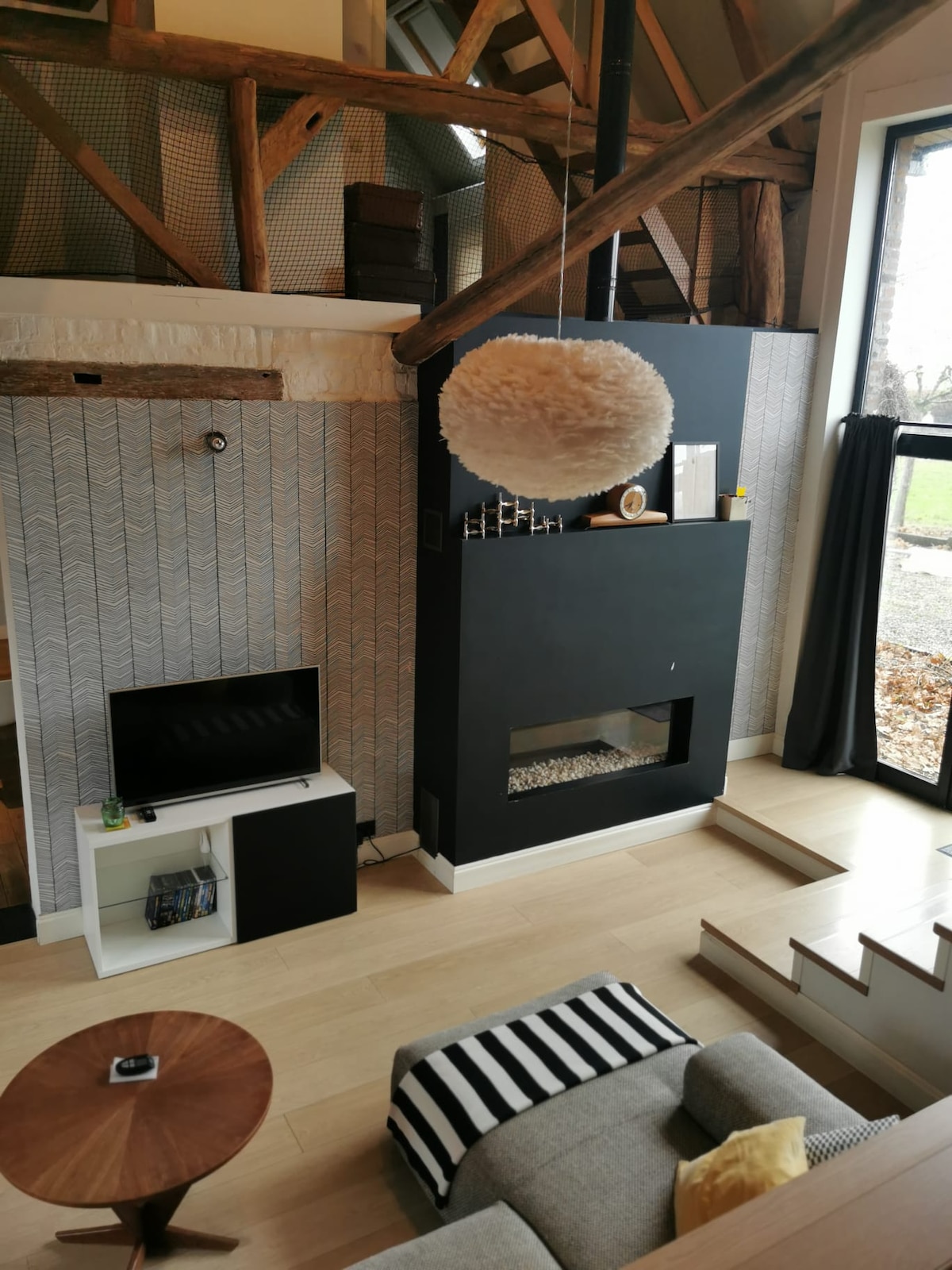 A cozy living area features a modern television mounted on a white unit beside a statement fireplace. A light-colored sofa is positioned nearby, complemented by a round wooden coffee table. Large windows allow natural light to brighten the space, showcasing elements of natural wood and neutral tones.