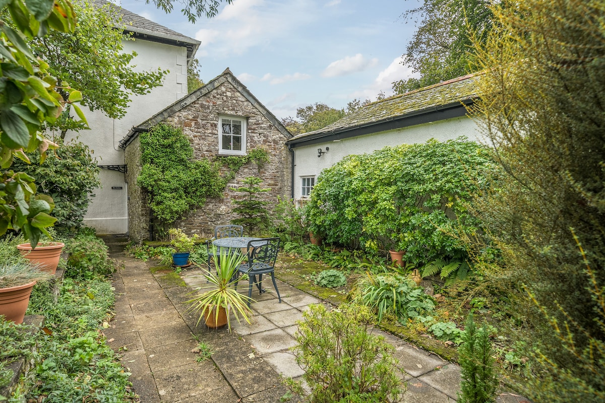 A charming garden area features a stone patio surrounded by lush greenery and potted plants. A table and chairs invite outdoor relaxation, while a mixture of shrubs and flowers adds to the inviting atmosphere. The cottage's exterior reflects a rustic character amidst the vibrant landscaping.