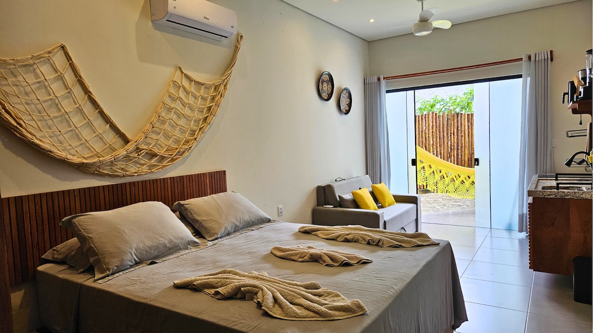 A spacious area combines the sleeping space with a comfortable sofa and kitchenette. The queen bed is dressed with neutral bedding, while the soft grey sofa features yellow accent cushions. Glass doors lead to a private outdoor area with a hammock, providing an inviting view.