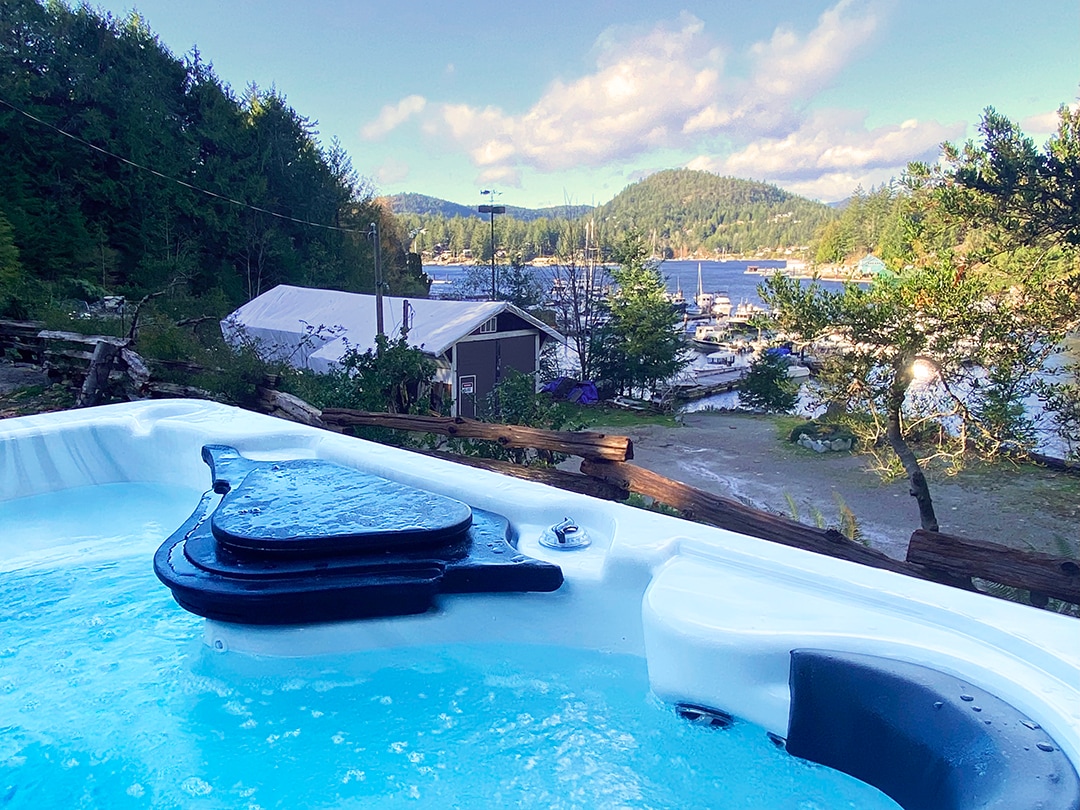 A hot tub is positioned on a wooden deck, showcasing bubbling water. In the background, the tranquil waterfront is visible, with boats docked amidst lush greenery and rolling hills, framed by a partly cloudy sky.
