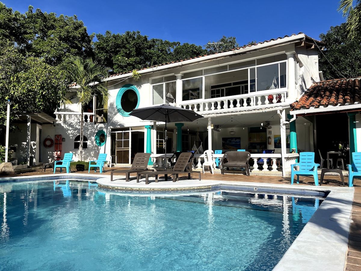 A large outdoor pool is surrounded by lounge chairs and patio furniture, with an umbrella providing shade. The two-story building features large windows and a balcony that overlook the pool area, framed by lush greenery.