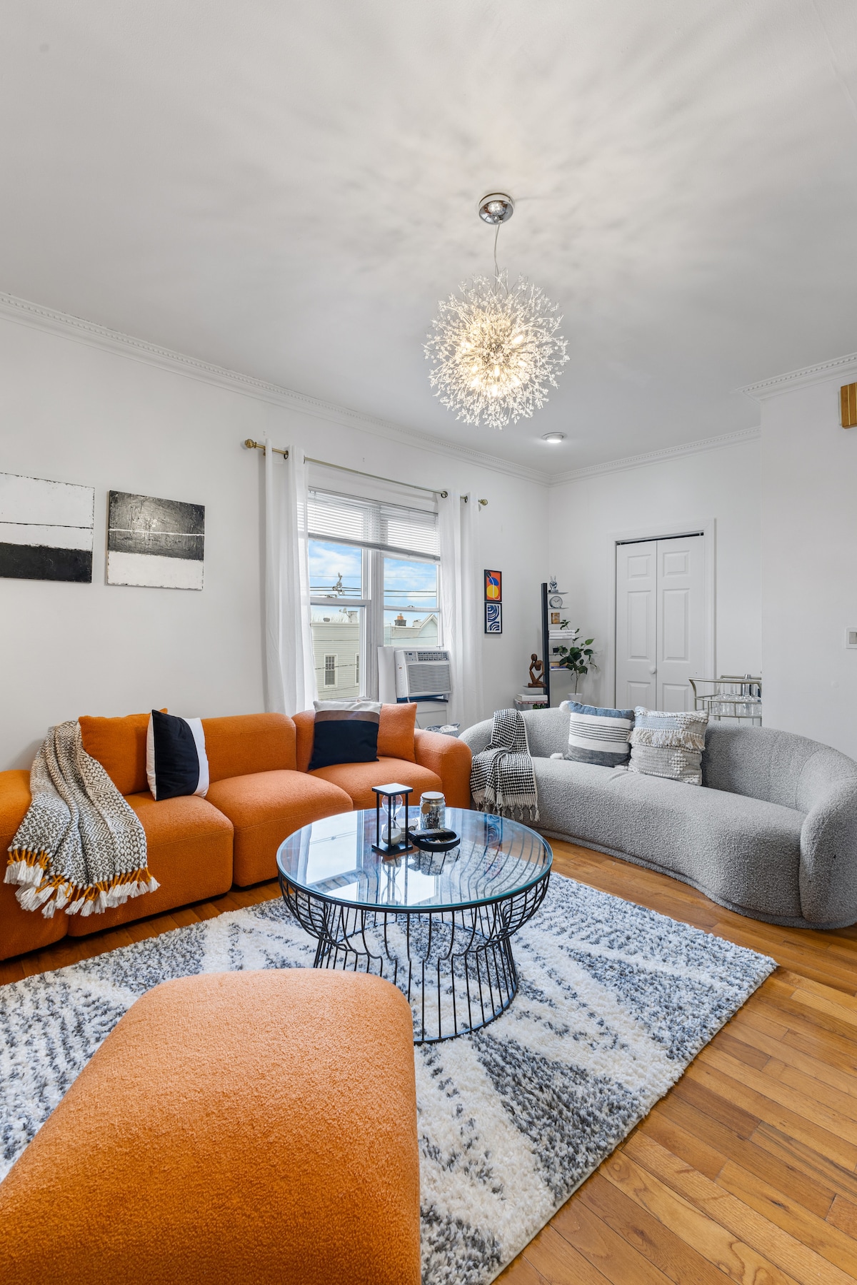 A modern living room features a large orange sofa and a soft gray sectional, arranged around a circular glass coffee table. Natural light enters through a window, illuminating the hardwood floor and a decorative area rug. A contemporary chandelier hangs from the ceiling, adding a stylish touch.