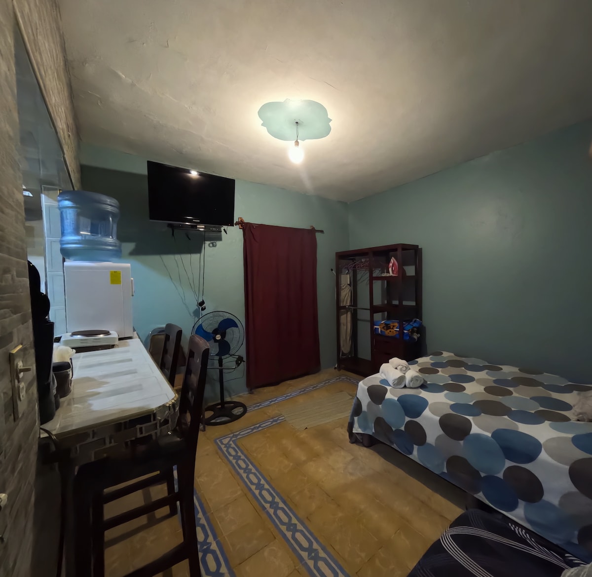 A compact room features a spacious bed with a polka dot bedspread and a wall-mounted television. A small kitchenette is located adjacent to a dining area with four chairs. A large ceiling fan is positioned in the corner, and a red curtain adds a pop of color.