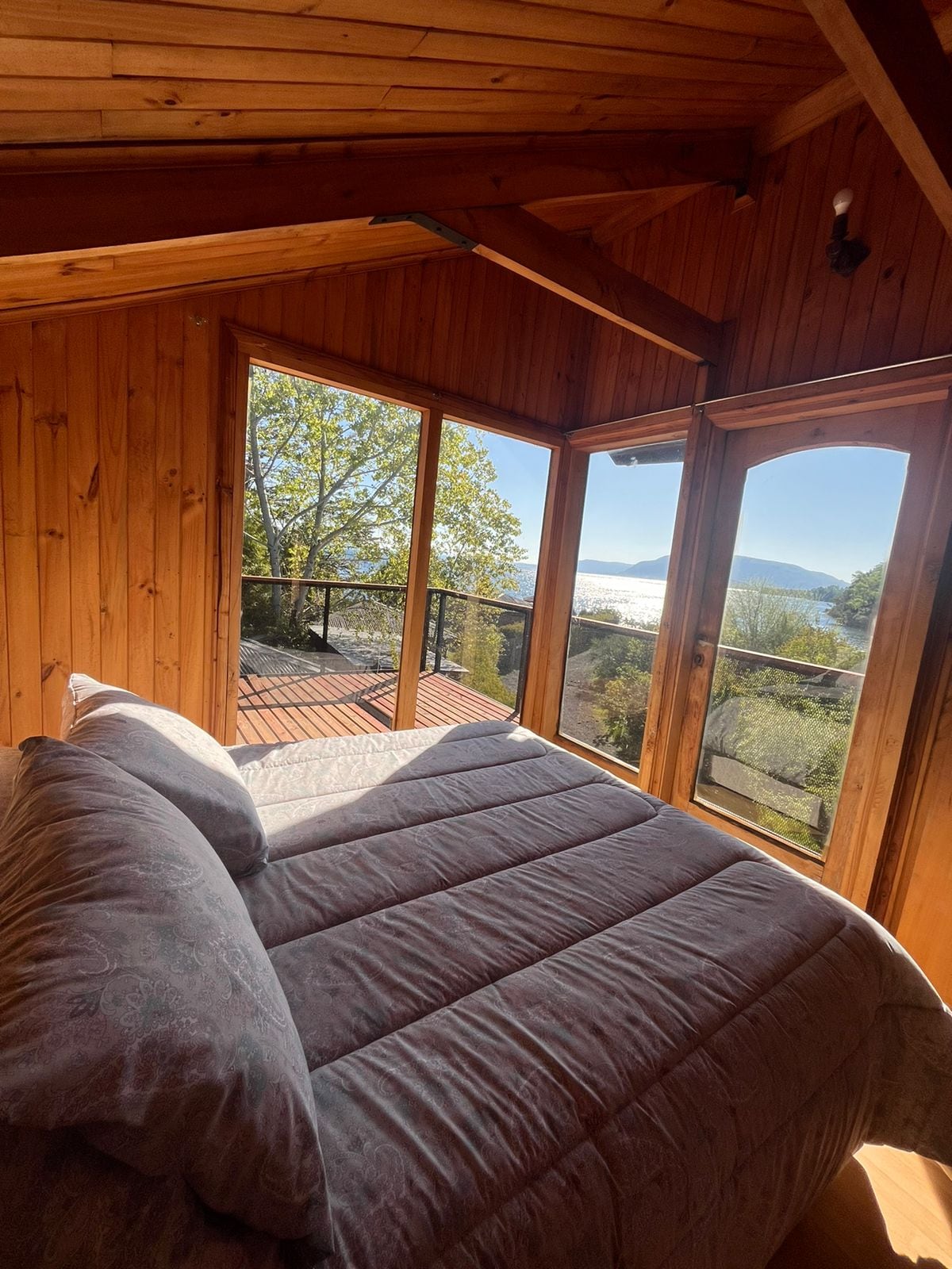A cozy sleeping area is framed by large windows, offering views of the lake and surrounding nature. Natural wooden walls complement the spacious bed dressed in a soft grey coverlet, with sunlight illuminating the peaceful setting.