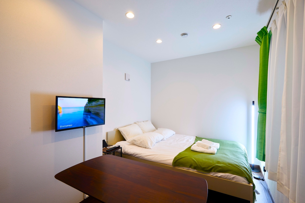 A well-lit sleeping area features a spacious bed with soft bedding and two pillows. A small table is visible in front of the bed, providing space for relaxation. Green curtains frame the window, and a mounted television adds convenience to the room.