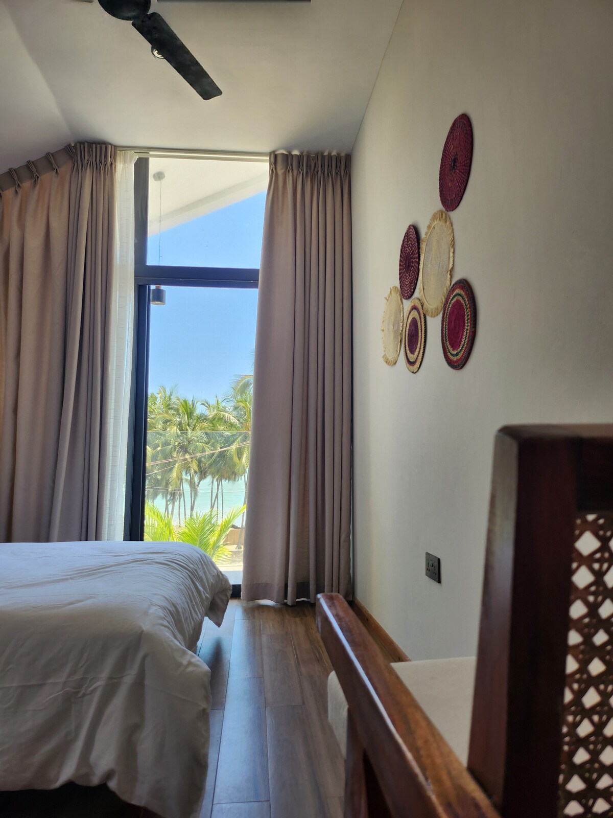 A cozy room features a spacious bed with crisp white linens and warm wooden flooring. Natural light enters through large glass doors, revealing a view of palm trees outside. Decorative wall hangings add a touch of texture to the calming atmosphere.