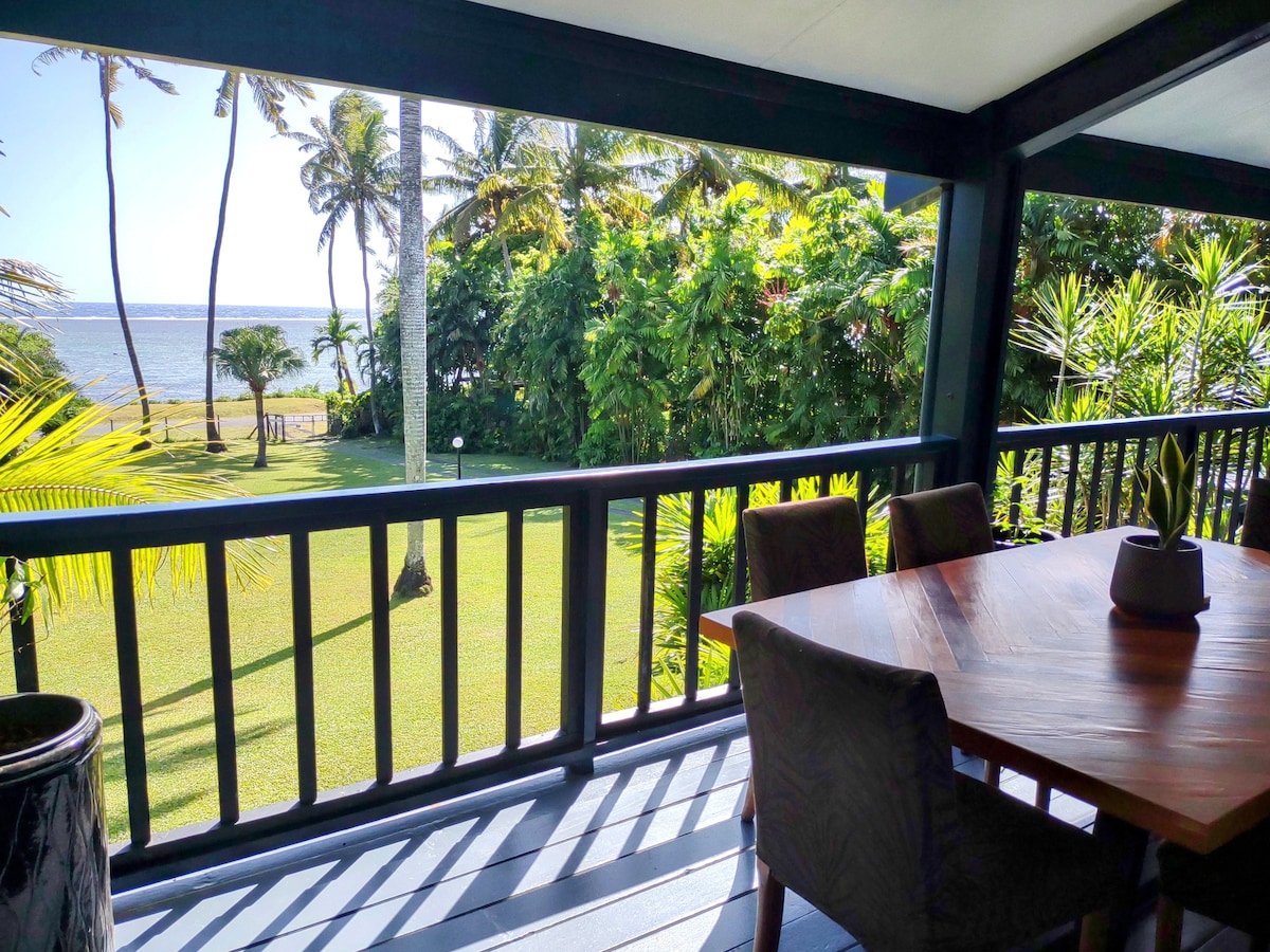 A spacious outdoor deck features a wooden dining table surrounded by chairs, overlooking a lush garden and ocean views. Tall palm trees and tropical plants frame the scene, creating a serene setting for relaxation and dining in the warm tropical climate.