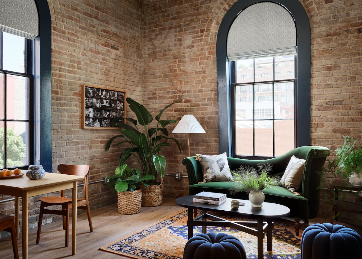 A cozy living space features exposed brick walls and large arched windows. A green sofa is complemented by a black coffee table and a wooden dining table. A potted plant adds a touch of nature, and a patterned area rug enhances the warmth of the room.