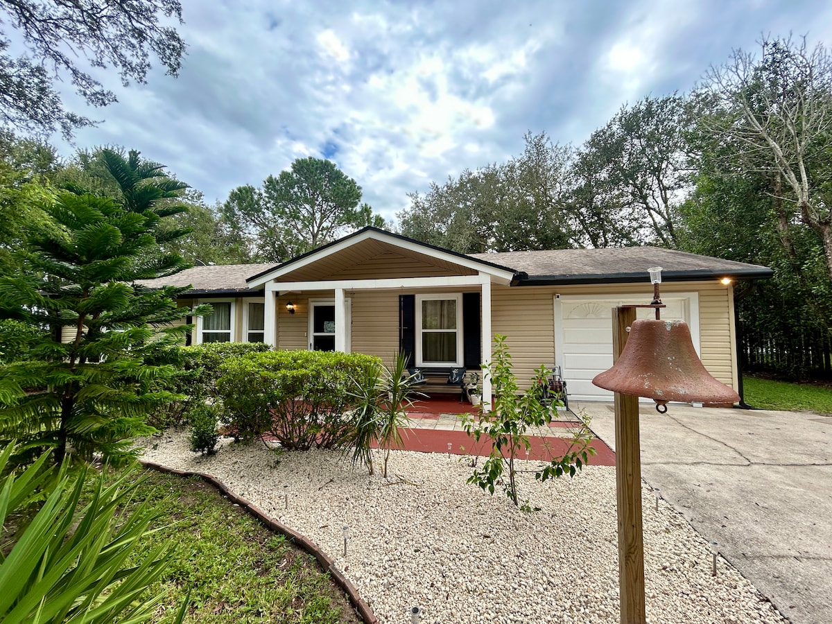A single-story home is surrounded by lush greenery, including a palm and decorative bushes. The welcoming front porch features white rocking chairs. A gentle bell stands in the foreground, while the driveway leads to a garage door on the right side.