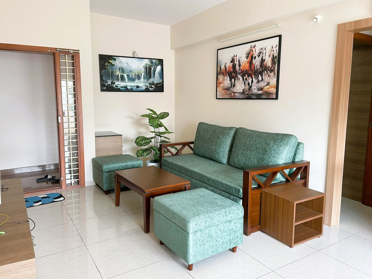 A cozy living area features a plush sofa, complemented by matching ottomans and a coffee table. Two framed artworks adorn the wall, adding a touch of character. Natural light reflects off the glossy tile floor, enhancing the spacious feel of the room.