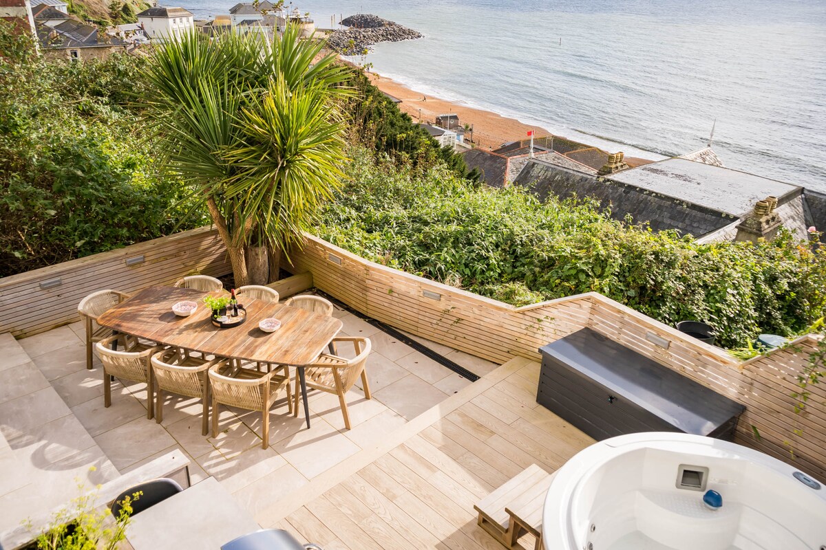 A private patio features a wooden table surrounded by eight chairs, ideal for alfresco dining. A barbecue grill and wooden hot tub are positioned nearby, with lush greenery framing the space. Stunning sea views overlooking the beach complete the tranquil outdoor setting.