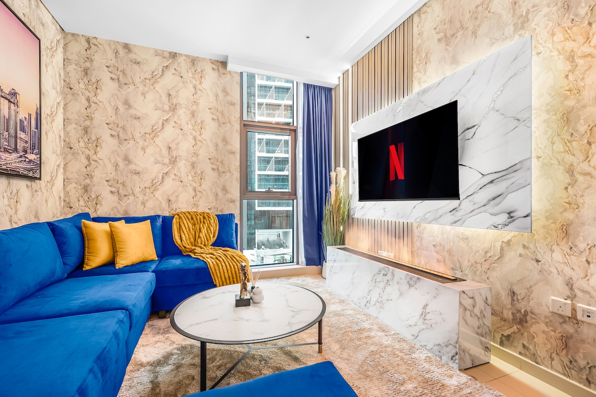 A modern living area features a large blue sectional sofa adorned with yellow cushions. A marble coffee table sits in front of the sofa. A flat-screen television is mounted on a stylish marble wall, with natural light streaming in from a nearby window.