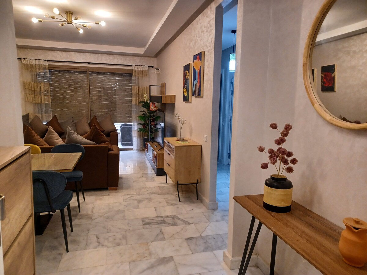 A contemporary living area features a mix of comfortable seating with cushions, a sleek coffee table, and a flat-screen television. Light-colored walls and marble flooring enhance the space, while soft natural light streams through large windows adorned with sheer curtains.