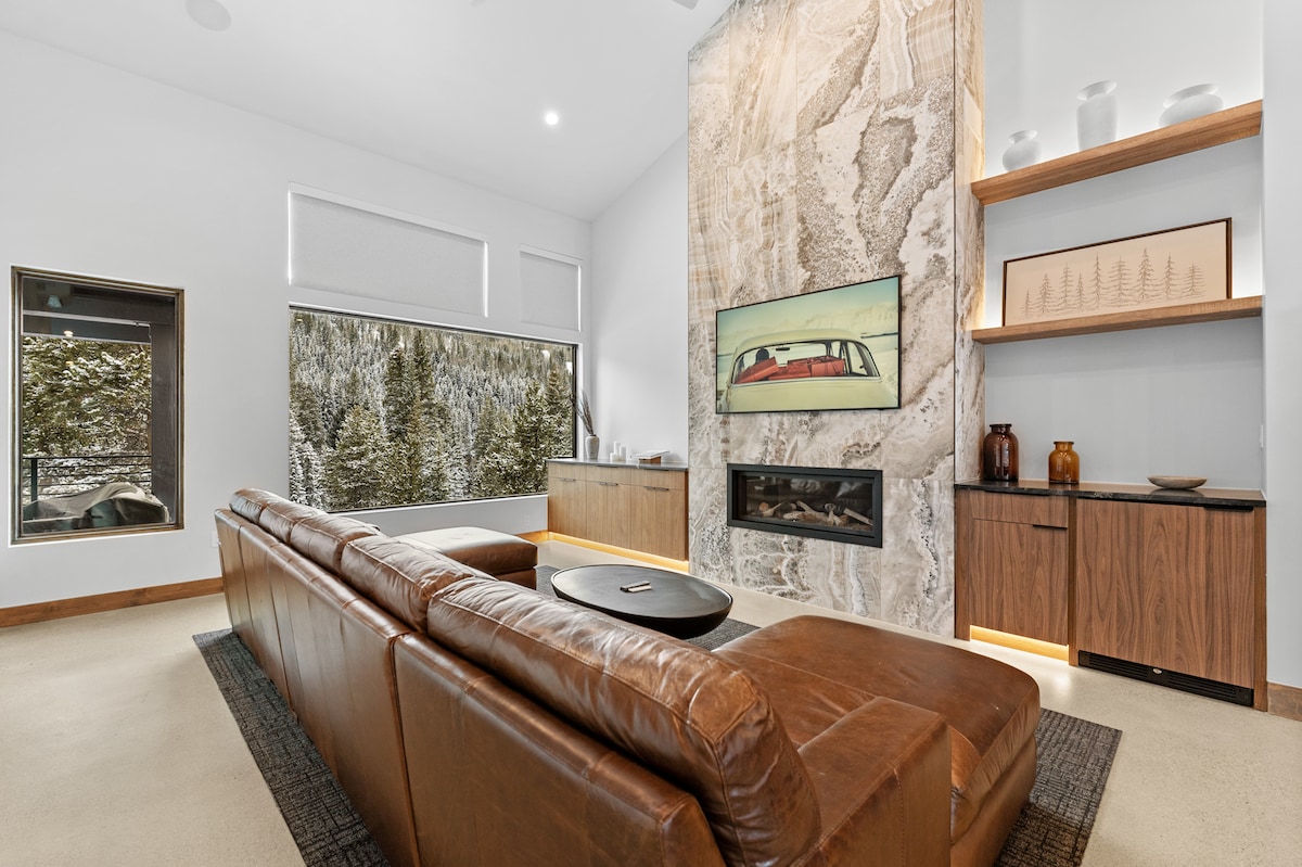 A spacious living area features a modern leather sofa arranged around a low table. A large stone fireplace serves as a focal point, complemented by a flat-screen TV mounted above. Oversized windows reveal views of surrounding trees, ensuring ample natural light.