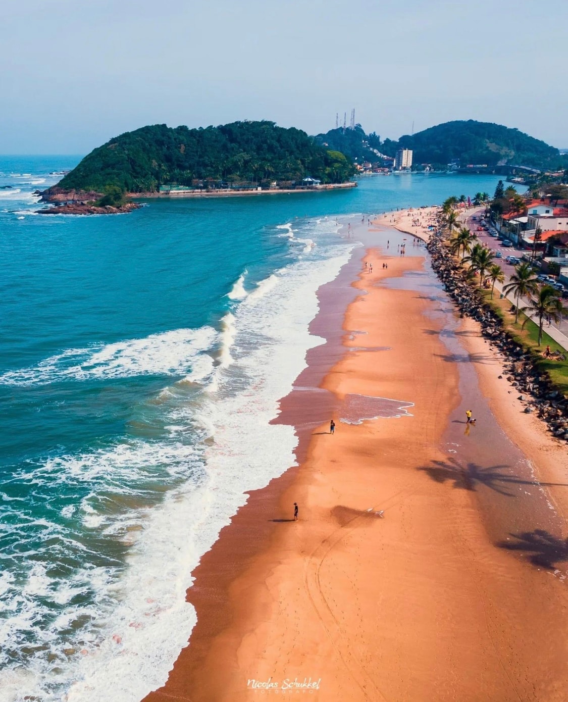 An aerial view showcases a sandy beach bordered by gentle waves lapping at the shore. Palm trees line the coastline along with a walking path, while distant hills rise in the background, and people can be seen enjoying the beach atmosphere.