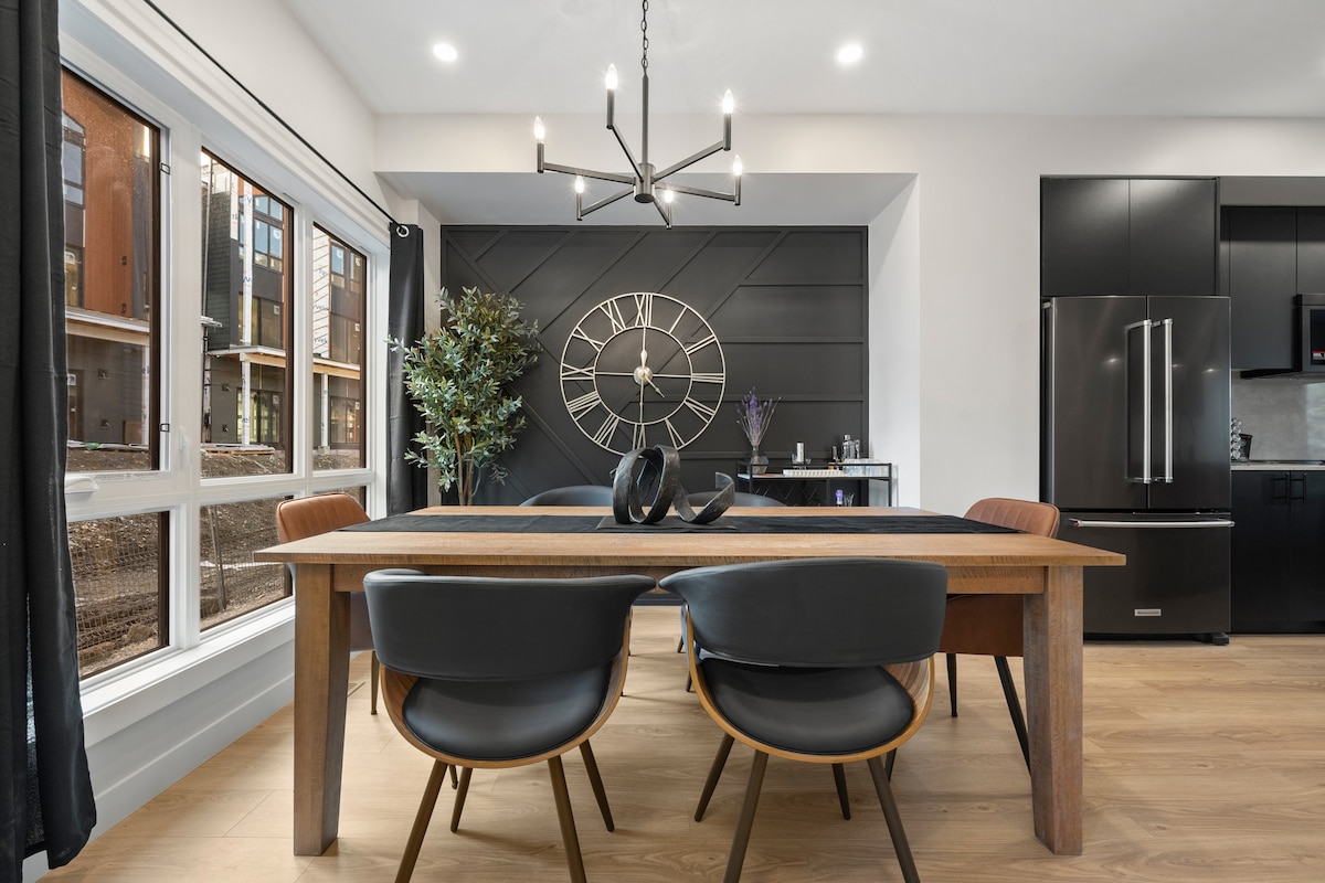 A dining area features a long wooden table surrounded by modern black chairs. A striking wall clock is mounted on a dark accent wall, while large windows provide natural light and views of the surroundings. Plants add a touch of greenery to the space.
