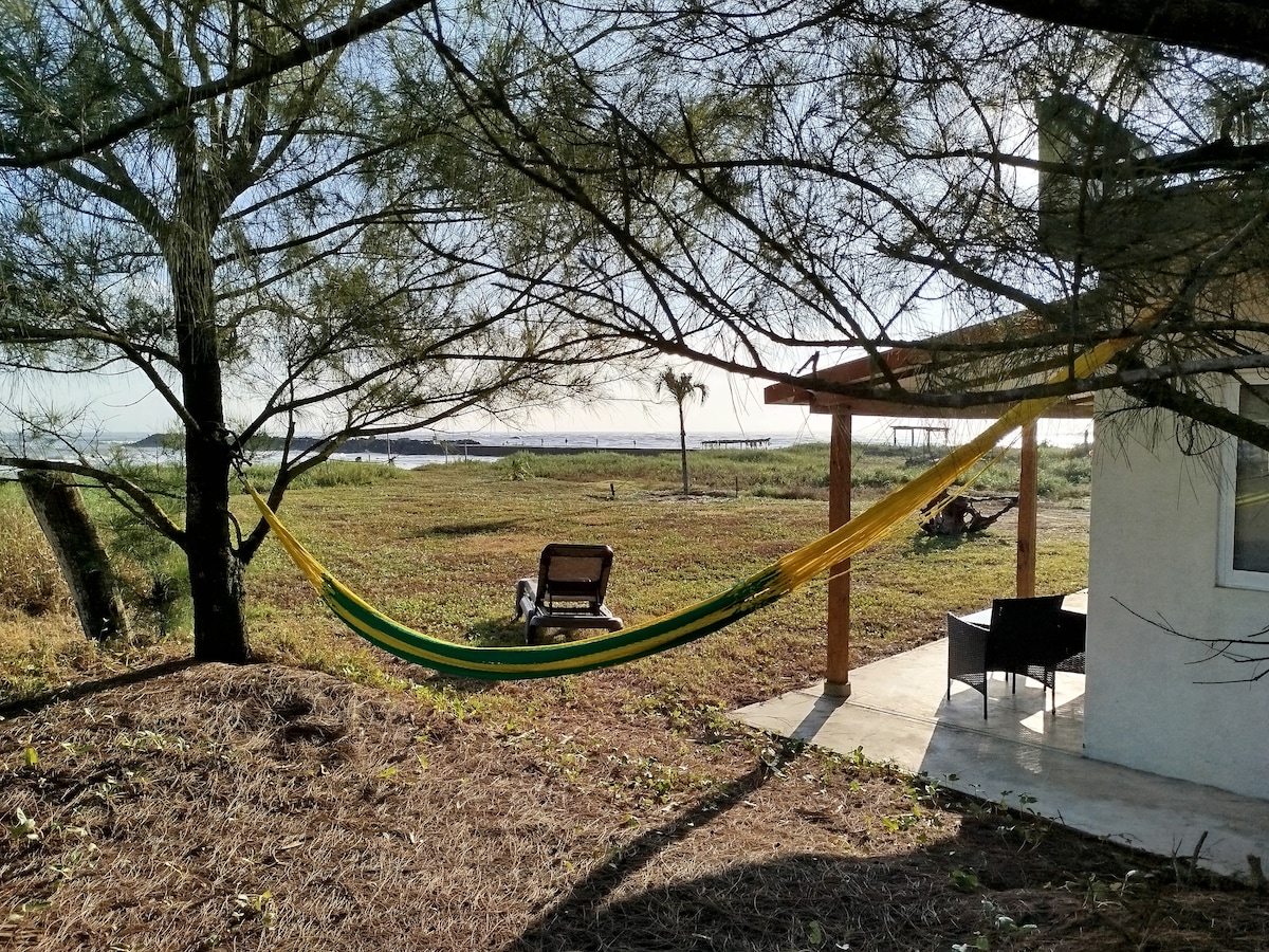 A vibrant hammock is suspended between two trees, inviting relaxation. Nearby, a comfortable chair is positioned under a wooden structure, partially shaded by foliage. The landscape features open grassy areas and a clear view of the water's edge, providing a peaceful outdoor setting.
