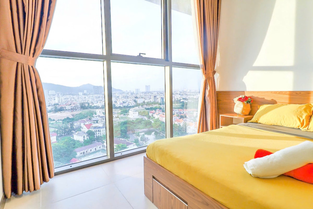 A bright bedroom is showcased with a large window offering expansive views of the cityscape. Soft yellow bedding is complemented by decorative pillows. Natural light filters through sheer curtains, enhancing the relaxing ambiance of the space.