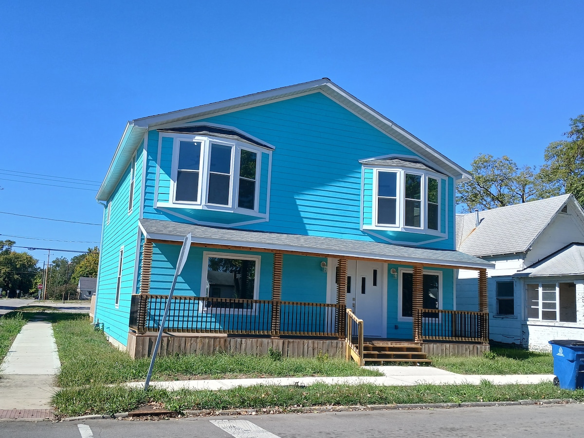 A bright blue two-story house is featured, showcasing large dual-pane picture windows that allow natural light. The house is set back from the street, offering a welcoming porch with steps leading to the front door. Lush green grass surrounds the base of the property.