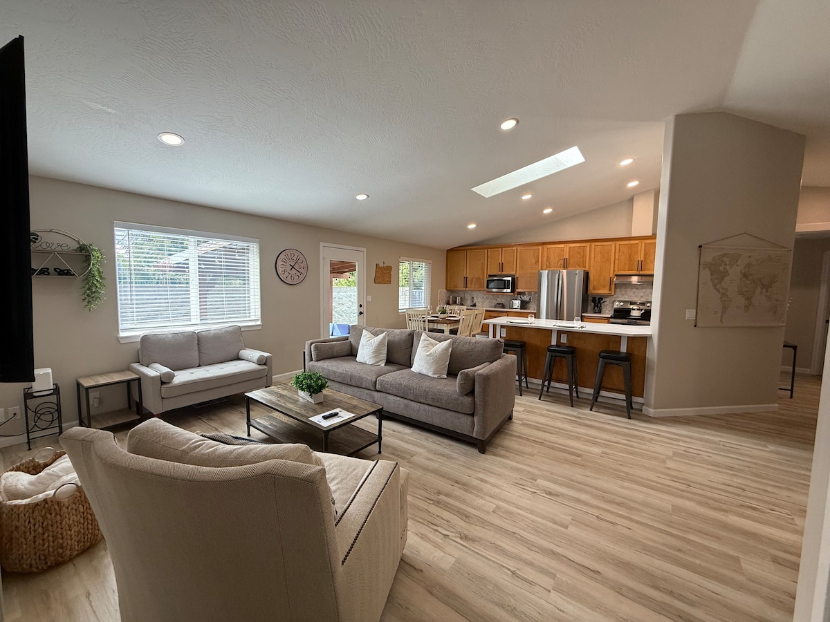 An open-concept living area features a spacious layout with comfortable seating arranged around a low coffee table. Natural light enters through large windows, illuminating the contemporary kitchen with wooden cabinetry and stainless steel appliances in the background.
