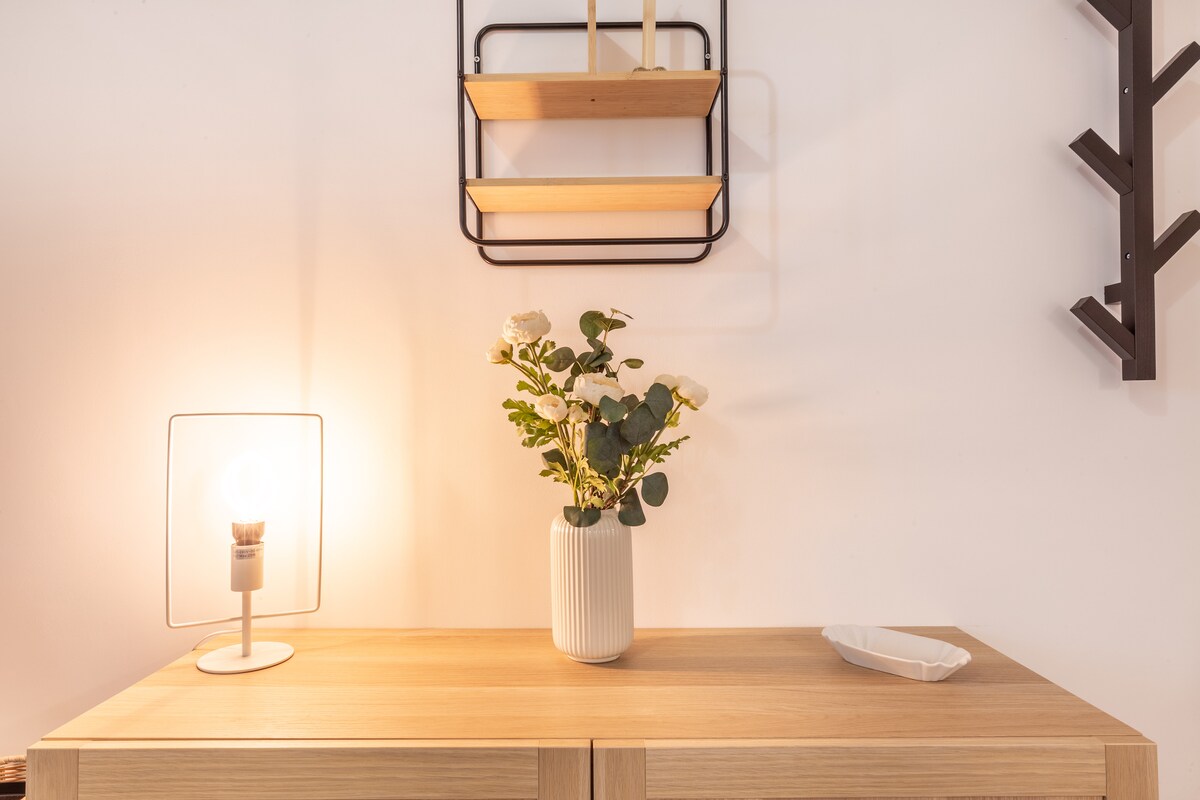 A wooden side table displays a minimalist arrangement featuring a white vase with fresh flowers, complemented by a small, elegant dish. A soft glow from a nearby lamp enhances the ambiance, while a simple wall shelf above holds decorative items.