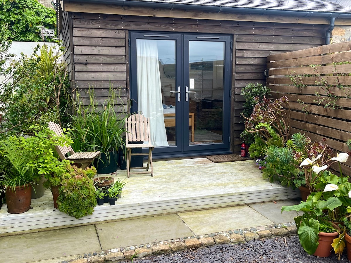 A compact outdoor area is featured, with two wooden chairs positioned on the wooden deck. Potted plants and greenery frame the entrance, leading to the sliding glass doors that provide access to the interior space. The area is well-maintained and inviting.