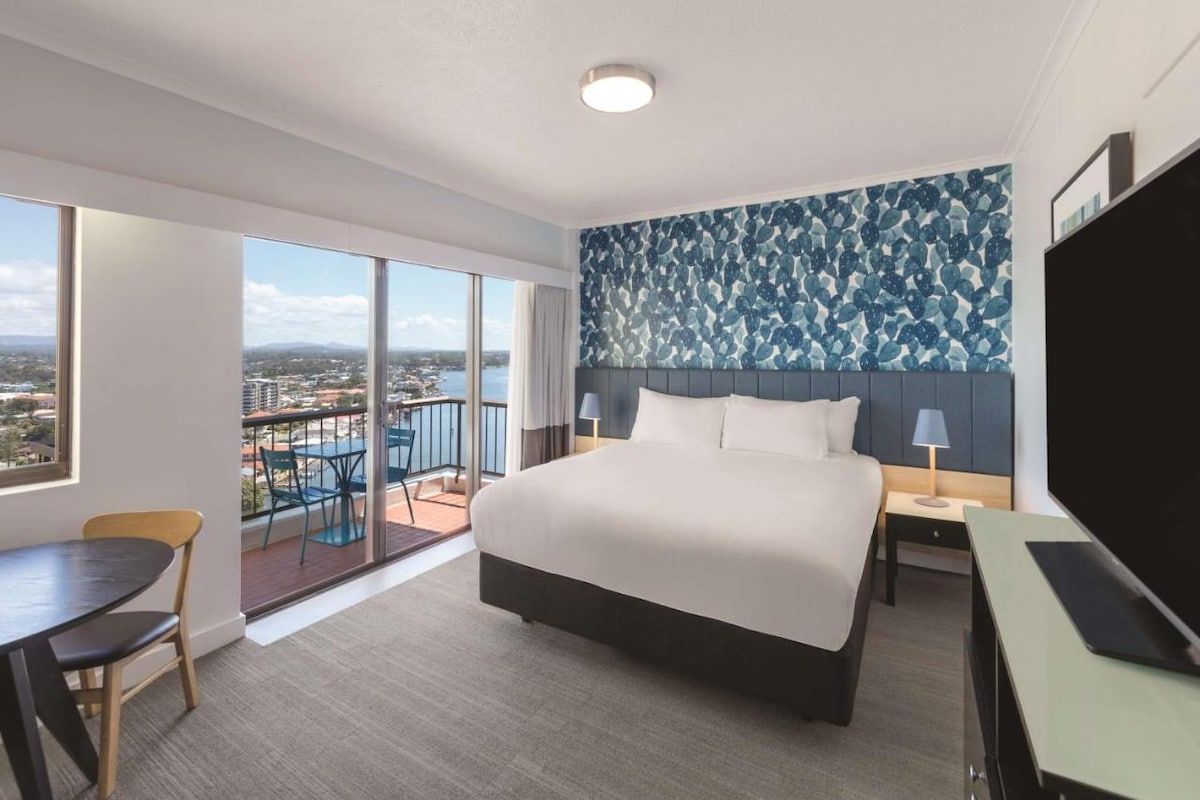 A bright and modern room features a spacious bed with a stylish blue accent wall. Large glass doors open to a balcony, revealing city views. A small dining table and TV add functionality to the space, complemented by soft natural light.