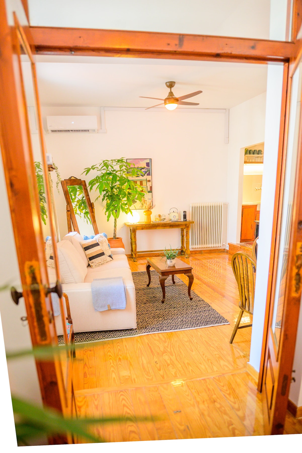 A welcoming living space is visible through an open door, featuring a light-colored sofa accompanied by decorative pillows. A wooden table sits in front, paired with a chair. Lush green plants are present, enhancing the relaxed ambiance, along with a modern ceiling fan overhead.