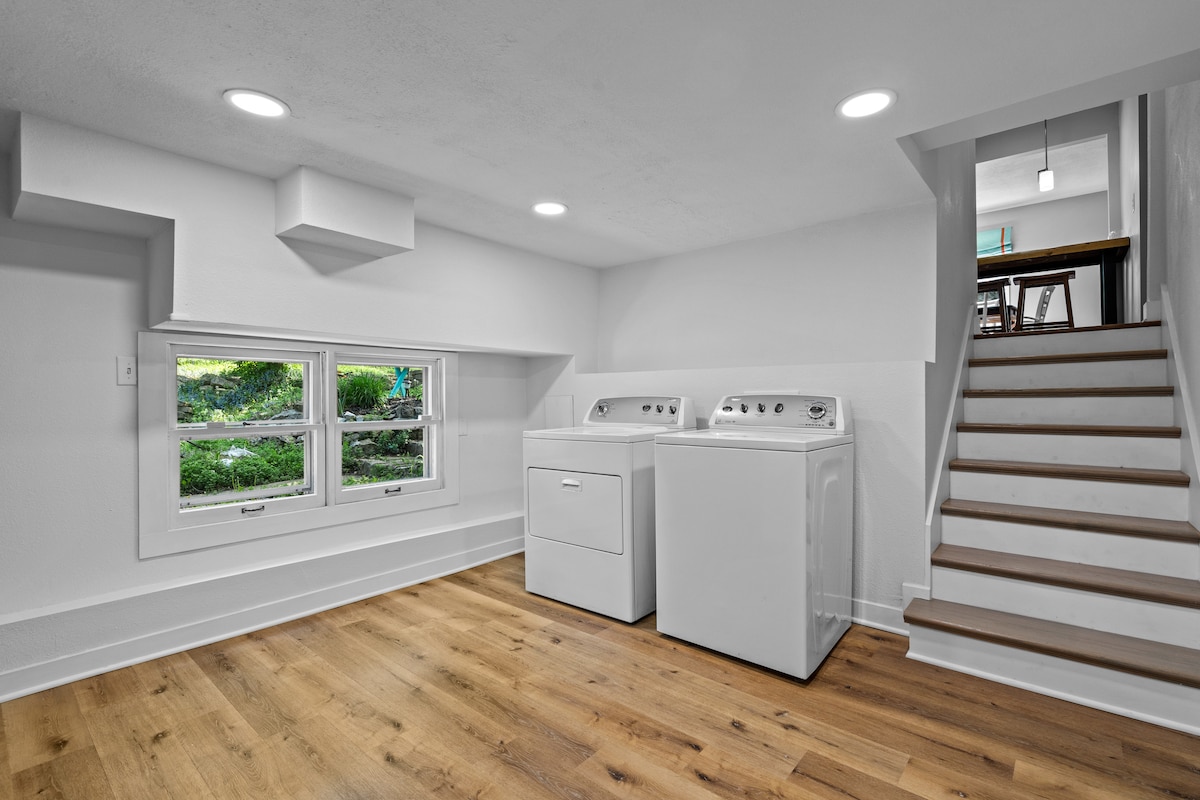 A well-lit laundry area features modern washing machines and a dryer against a light-colored wall. A set of steps leads up to the main floor, while ample natural light enters through a set of windows, showcasing a view of the greenery outside.