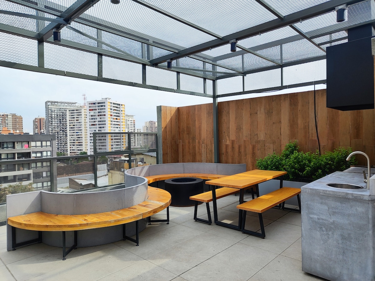 A spacious rooftop area is equipped with concrete seating and wooden tables. A built-in kitchen space with a sink is visible. The surrounding area boasts a wooden privacy fence, and city views are seen in the background.