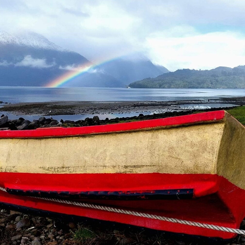 A colorful boat is partially buried in pebbles at the water's edge. A vibrant rainbow arcs across the sky above a serene body of water, flanked by green hills and distant mountains. Soft clouds create a tranquil atmosphere in the background.