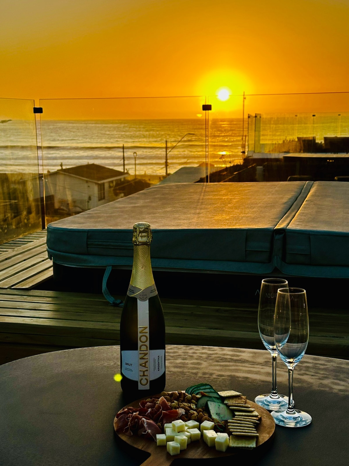 A rooftop terrace is set for relaxation as the sunset casts a warm glow over the ocean. A bottle of champagne stands alongside two glasses, beside a wooden platter featuring assorted cheeses and cured meats. The scene overlooks the beach and surrounding seaside structures.