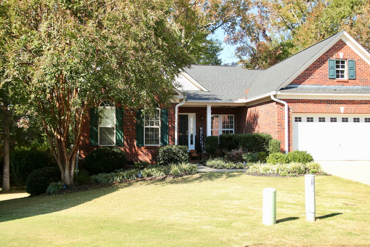 The exterior of a red brick home is showcased, complemented by green shutters and a well-maintained front yard. The entrance features a covered porch, surrounded by shrubs and trees, contributing to a welcoming and serene ambiance in a quiet neighborhood.