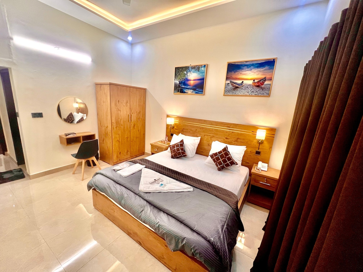 A spacious sleeping area features a large bed with neutral-toned bedding. Two framed artworks hang above the headboard. Nightstands with lamps are positioned on either side of the bed, and a chair and mirror are placed against the wall in an organized layout.