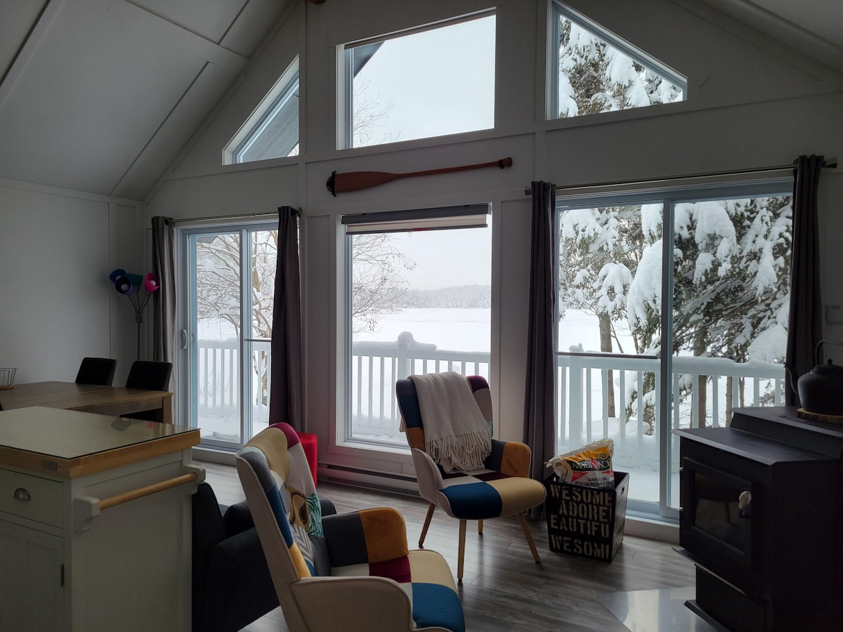 Large windows provide a panoramic view of the snow-covered surroundings and lake. A cozy seating area includes colorful chairs and throws. A small fireplace adds warmth, while a kitchen area with a stone countertop complements the spacious design of the living area.