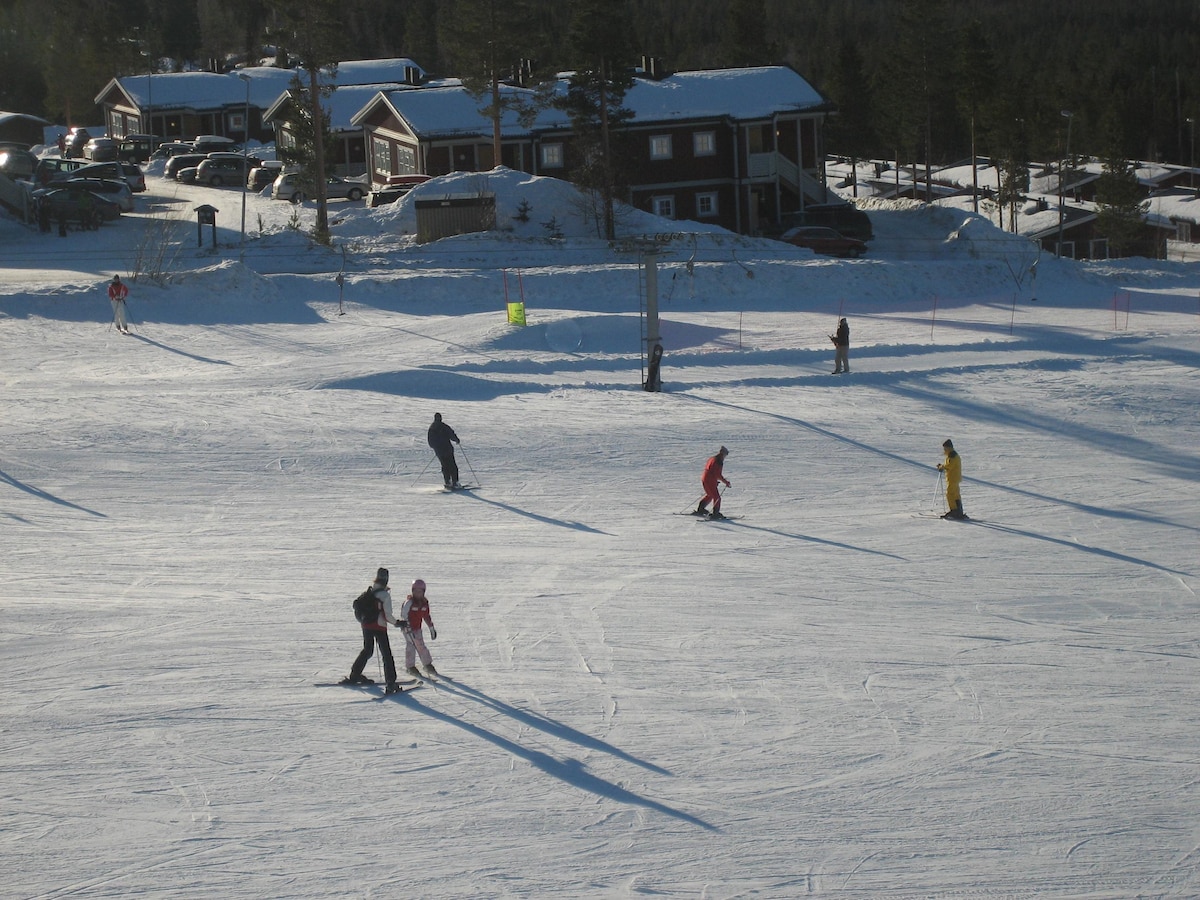 Several skiers are seen navigating a wide snowy slope, showcasing a variety of ski attire. In the background, a cluster of brown wooden lodges is visible, set against a backdrop of evergreen trees under a clear blue sky. The tranquil winter scene conveys a sense of outdoor activity.