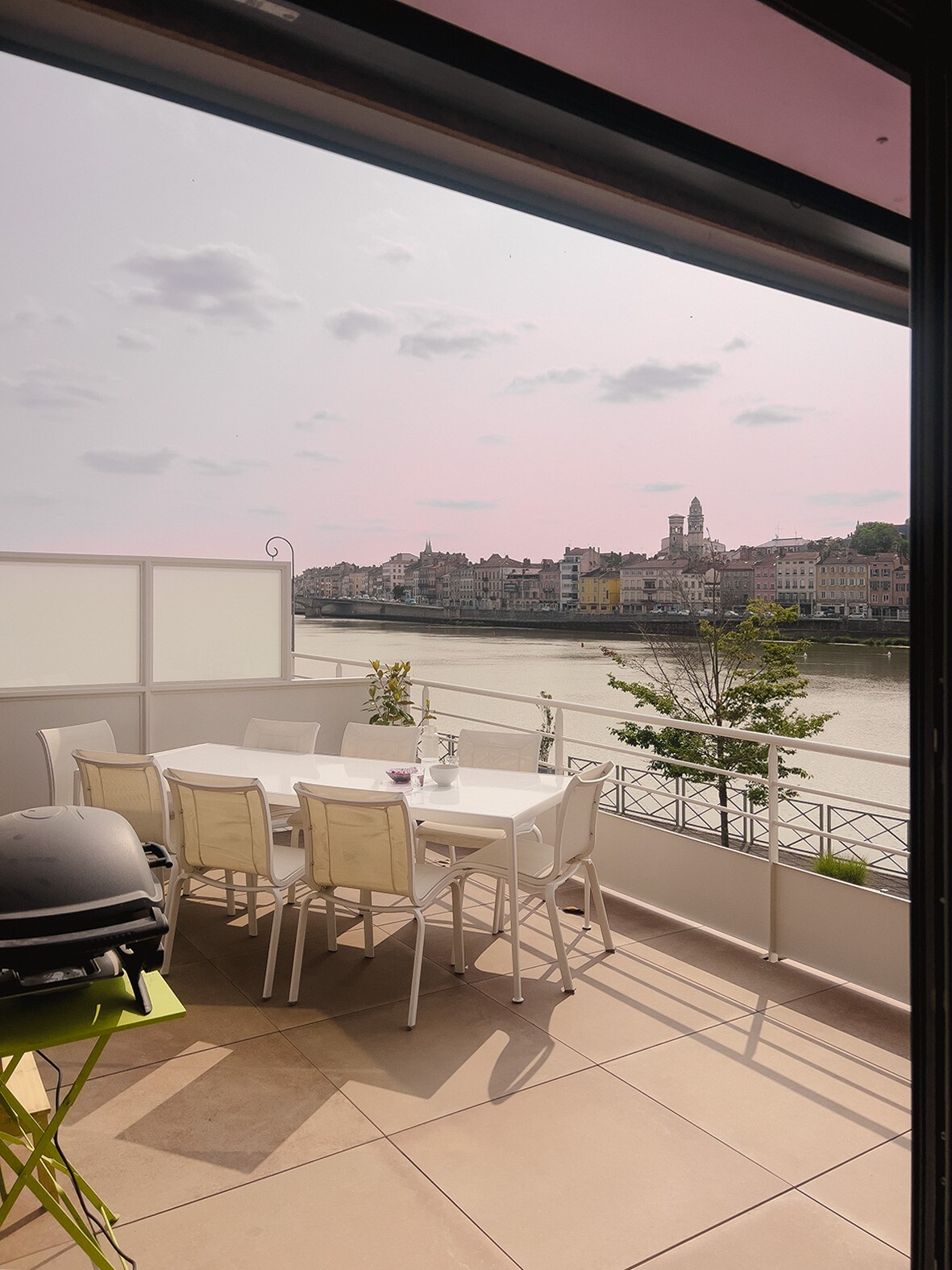 A terrace area features a white dining table surrounded by several light-colored chairs, overlooking a river and a scenic view of nearby buildings. A barbecue grill is positioned in the corner, and a few green plants add a touch of nature to the space.